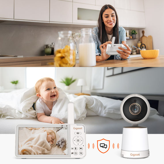 Mère assise à la table de la cuisine avec un moniteur, bébé souriant dans son lit. Communication bidirectionnelle via Gigaset BABY 500 Video.