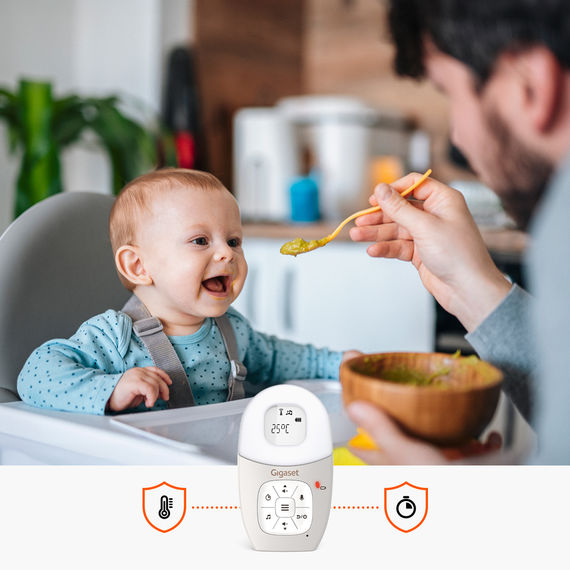 Babyphone mit Elterneinheit. Im Hintergrund ein Baby im Hochstuhl, das von seinem Vater gefüttert wird.