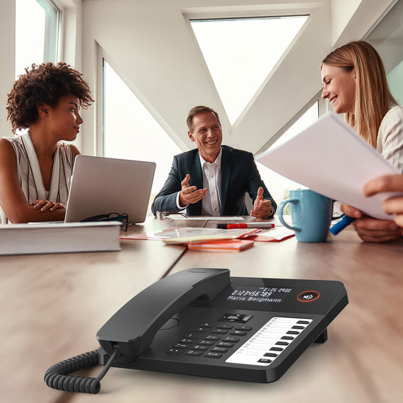 Moderno teléfono de sobremesa con pantalla y teclas de función, al fondo un grupo en una reunión.