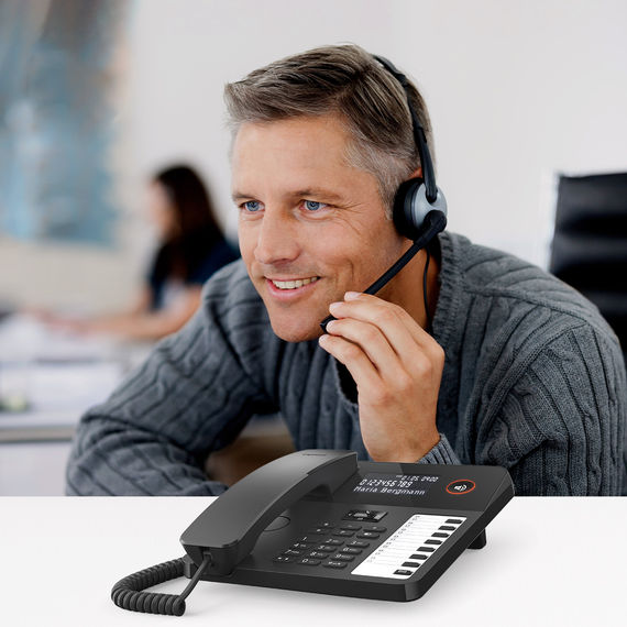 Modern desk phone with display and function keys, in the background a man with a headset.