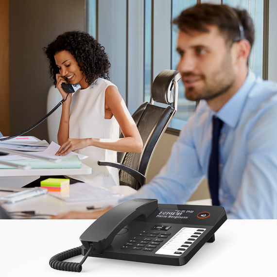 Moderno teléfono de sobremesa con pantalla y teclas de función, al fondo un hombre con auriculares y una mujer al teléfono.